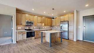 Open-concept apartment kitchen with a center island, wood cabinetry, stainless steel appliances, and wood-look flooring flowing into the adjacent living area.