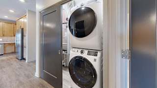 Apartment laundry closet with a stacked front-load washer and dryer, conveniently located off the kitchen with wood-look flooring and modern interior finishes.