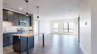 a modern kitchen with a table island and dark cabinets next to a living room with two large windows