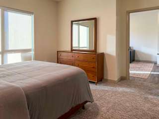 Bright bedroom with carpet flooring, large window, bed, and wood dresser with mirror, opening to an adjacent living area.