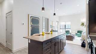 A kitchen with dark wooden cabinets and a large island, in front of a spacious living room with floor-to-ceiling windows.
