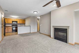 Open-concept apartment living room with carpet, a corner fireplace, ceiling fan, and breakfast bar overlooking a kitchen with wood cabinets and black appliances.