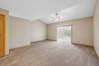 Spacious, empty apartment living room with vaulted ceilings, neutral carpet, a ceiling fan, and a large sliding glass door with vertical blinds that fills the space with natural light.