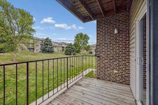 Covered apartment balcony with wood decking, a brick accent wall, metal railing, and views of the landscaped grounds and neighboring apartment buildings.