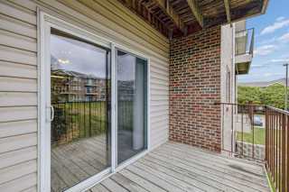 Covered apartment balcony with wood decking, a sliding glass door, brick accent wall, metal railing, and views of the surrounding apartment community and green space.