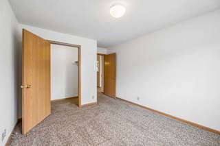 Bright, empty apartment bedroom with neutral carpet, white walls, wood-trimmed doors, and an open closet area leading to the adjoining hallway.