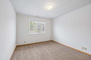 Bright, empty apartment bedroom with neutral carpet, white walls, wood trim, and a single window with blinds providing natural light.