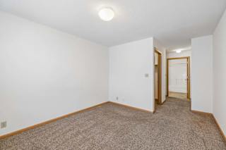 Bright, empty apartment bedroom with neutral carpet, white walls, and an open layout leading to the hallway and adjoining bathroom.