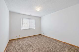 Bright, empty apartment bedroom with neutral carpet, white walls, wood trim, and a window with blinds that provides natural light.