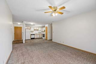 Spacious apartment living room with neutral carpet, a ceiling fan, and an open layout leading to the kitchen with a breakfast bar, wood-tone cabinets, and white appliances.