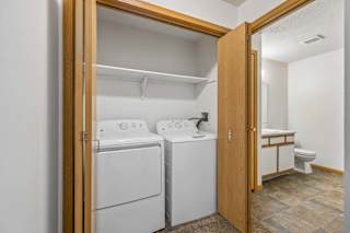 Apartment laundry closet with a full-size washer and dryer, bi-fold wood doors, and an adjacent bathroom featuring a single-sink vanity and tub/shower combination.