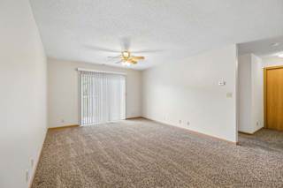 Spacious apartment living room with neutral carpet, a ceiling fan, vertical blinds over a sliding glass door, and an open layout leading to the hallway.
