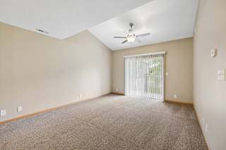 Spacious apartment living room with carpeted flooring, a vaulted ceiling, a ceiling fan, and a large sliding glass door with vertical blinds leading to a private patio or balcony.