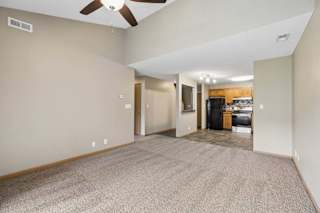 Open-concept apartment living room with carpeted flooring, a ceiling fan, vaulted ceilings, and a view into the kitchen featuring wood cabinetry and black appliances.