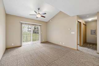 Spacious apartment living room with carpeted flooring, a vaulted ceiling, a ceiling fan, and a large sliding glass door with vertical blinds providing natural light and access to a private patio or balcony.