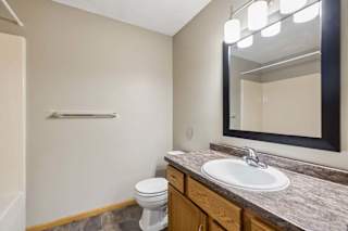 Apartment bathroom with a single-sink vanity, wood cabinetry, a granite-look countertop, a framed mirror with modern lighting, and a tub-shower combination with a white surround.