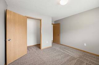 Bright apartment bedroom with carpeted flooring, an open closet with sliding doors, neutral walls, and a separate entry door for convenient access.