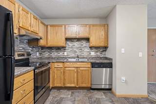 Apartment kitchen with wood cabinetry, a tile backsplash, laminate countertops, black and stainless steel appliances, and a functional layout adjacent to the entryway.