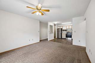 Spacious open-concept apartment living room with carpeted flooring, a ceiling fan, and a view into the kitchen featuring white and wood-tone cabinetry, stainless steel and black appliances, and a functional open layout.