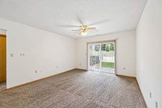 Spacious apartment living room with carpeted flooring, a ceiling fan, and a large sliding glass door with vertical blinds providing natural light and access to a private patio or balcony.