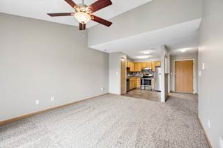 A room with a ceiling fan and carpeted floor and a kitchen in the background.