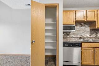 A kitchen with wooden cabinets. The pantry door is open. There is a grey tile backsplash and stainless steel appliances.