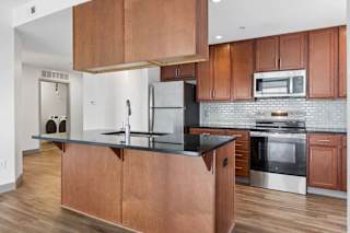 Apartment kitchen with wood cabinetry, black countertops, stainless steel appliances, tile backsplash, and visible laundry area.