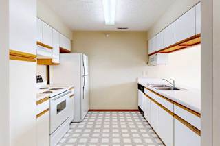 A galley kitchen with white cabinets with oak trim, white appliances, a double-basin sink, and a pass-through window countertop.