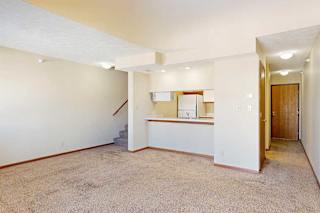 A carpeted living room with access to the kitchen via a pass-through window countertop, and a staircase to the upper loft area.