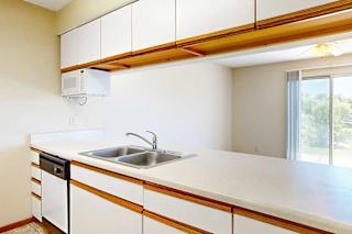 A galley kitchen featuring white cabinets with oak trim, a double basin sink, a mounted microwave, and a passthrough countertop window.