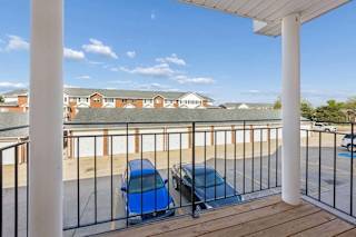 Covered apartment balcony with wood decking, white support columns, metal railing, and views of the community parking area and neighboring apartment buildings under a clear sky.