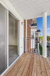 Covered apartment balcony with wood decking, a sliding glass door, brick accents, white support columns, and views of the surrounding apartment community.