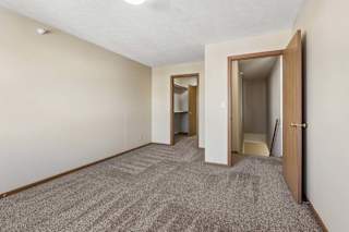 Spacious apartment bedroom with neutral carpet, beige walls, wood-trimmed doors, and an open layout leading to the hallway and closet area.