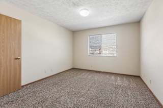 Bright, empty apartment bedroom with neutral carpet, beige walls, wood trim, and a window with blinds providing natural light.