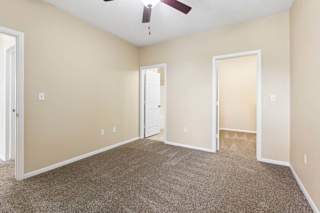 Carpeted apartment bedroom with beige walls, ceiling fan, and open doorways to nearby rooms.