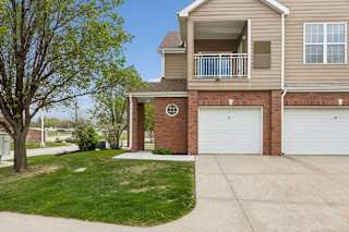 Apartment exterior with brick accents, tan siding, attached garages, balcony, driveway, and grassy lawn.