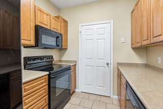 Galley-style apartment kitchen with wood cabinets, black appliances, oven, microwave, and tile floor.