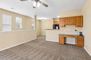 Open apartment living area with carpet, built-in desk cabinetry, wood cabinets, and windows with blinds.
