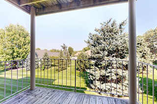 Apartment balcony with wood decking and metal railing overlooking a landscaped courtyard with mature trees and green lawn.