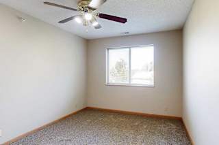 Bright bedroom with neutral walls, plush carpet, ceiling fan with light, and window for natural light.