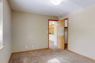 Carpeted apartment bedroom with neutral paint, ceiling light fixture, wood-trimmed door, and view into adjoining living area.