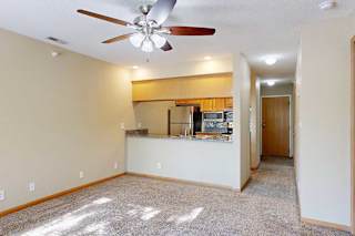 Open-concept living and dining area with ceiling fan, plush carpet, breakfast bar, and kitchen with wood cabinetry and stainless-steel appliances.