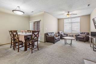 Spacious apartment living and dining area with neutral carpet, large window with blinds, ceiling fan, and sliding glass door leading to a patio.