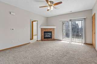 Bright, empty apartment living room with beige carpet, tiled fireplace, ceiling fan, and sliding glass doors opening to a balcony.