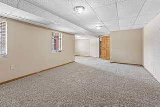 Spacious, empty lower-level room with neutral carpet, drop ceiling, recessed lighting, windows with blinds, and white brick wall.