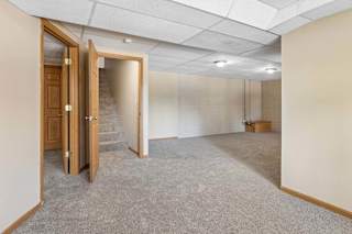 Empty lower-level room with neutral carpet, drop ceiling, white brick walls, overhead lighting, and stair access.