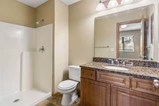 Bathroom with walk-in shower, wood vanity, speckled countertop, large mirror, and bright vanity lighting.