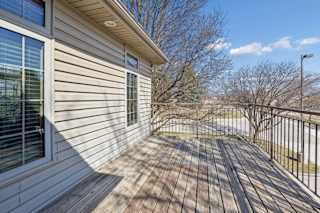 Spacious outdoor deck with wood plank flooring, metal railing, exterior siding, and views of trees and the community drive.