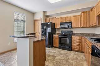 Kitchen with wood cabinetry, speckled countertops, black appliances, breakfast bar, tile flooring, and a large window.