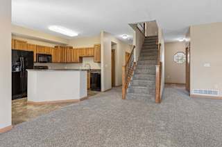 Open living area with neutral carpet, central staircase, kitchen with wood cabinetry, breakfast bar, and black appliances.